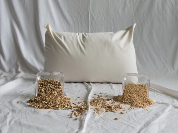 Dinkel-Hirse-Kopfkissen von Kissenkorner auf weißem Bettlaken. Vor dem Kissen steht eine Schüssel mit Dinkelspelzen und Hirseschalen.