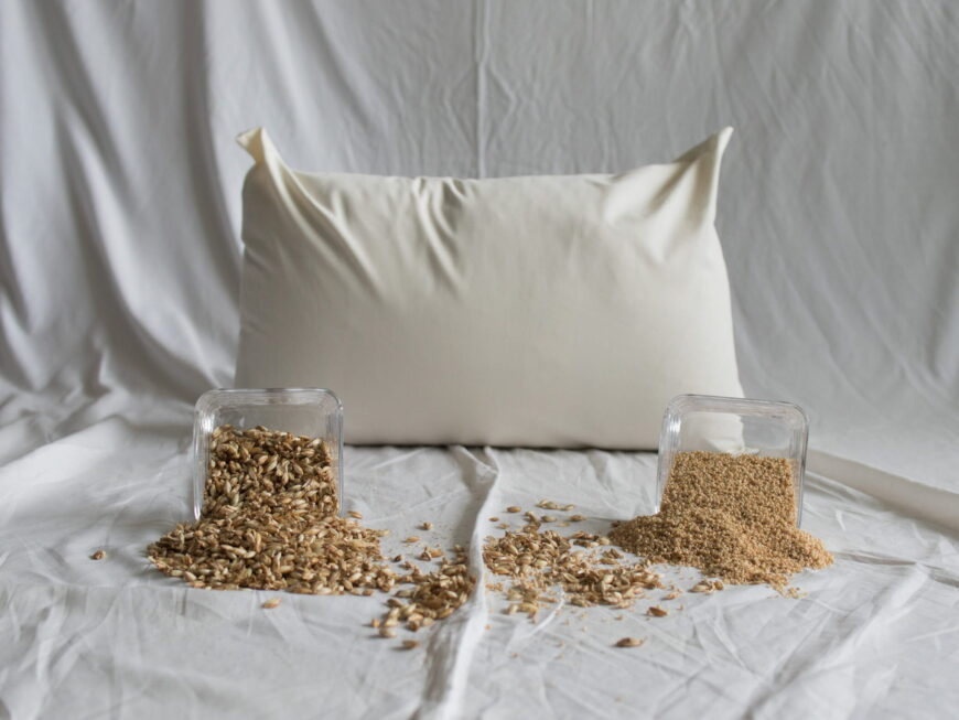 Dinkel-Hirse-Kopfkissen von Kissenkorner auf weißem Bettlaken. Vor dem Kissen steht eine Schüssel mit Dinkelspelzen und Hirseschalen.