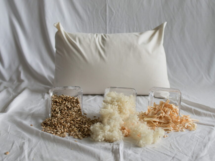 Dinkel-Schaf-Zirbe-Kopfkissen von Kissenkorner auf weißem Bettlaken. Vor dem Kissen steht eine Schüssel mit Dinkelspelzen und Schafwolle und Zirbenspänen.