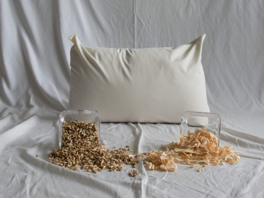 Dinkel-Zirbe-Kopfkissen von Kissenkorner auf weißem Bettlaken. Vor dem Kissen steht eine Schüssel mit Dinkelspelzen und Zirbenspänen.