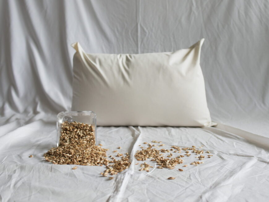 Dinkelkopfkissen von Kissenkorner auf weißem Bettlaken. Vor dem Kissen steht eine Schüssel mit Dinkelspelzen.