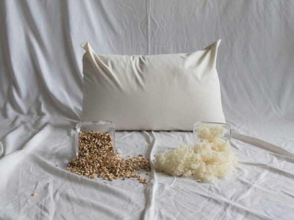 Dinkelspelzen-Schafwolle-Kopfkissen von Kissenkorner auf weißem Bettlaken. Vor dem Kissen steht eine Schüssel mit Dinkelspelzen und Schafwolle.