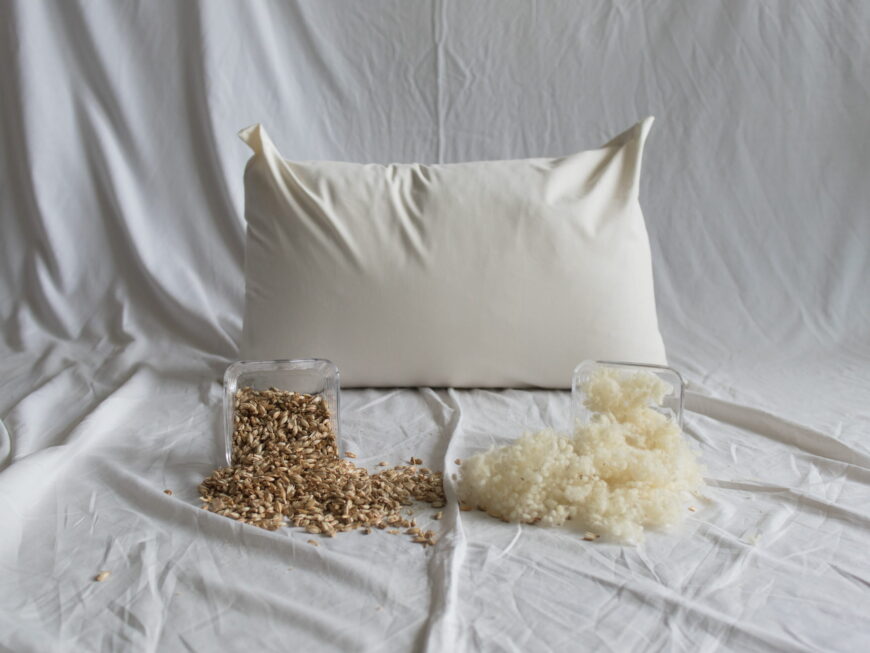 Dinkelspelzen-Schafwolle-Kopfkissen von Kissenkorner auf weißem Bettlaken. Vor dem Kissen steht eine Schüssel mit Dinkelspelzen und Schafwolle.