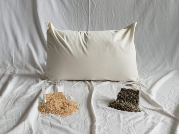 Hires-Kräuter-Kopfkissen von Kissenkorner auf weißem Bettlaken. Vor dem Kissen steht eine Schüssel mit Hirseschalen und einer 6 Kräutermischung.