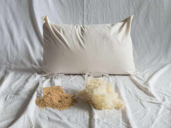 Hirse-Schafwolle-Kopfkissen von Kissenkorner auf weißem Bettlaken. Vor dem Kissen steht eine Schüssel mit Hirseschalen und Schafwolle.