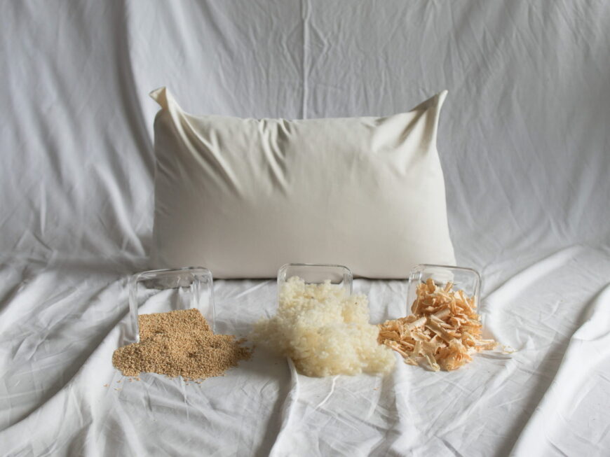 Hirse-Schafwolle-Zirbe-Kopfkissen von Kissenkorner auf weißem Bettlaken. Vor dem Kissen steht eine Schüssel mit Hirseschalen , Schafwolle und Zirbenspänen.