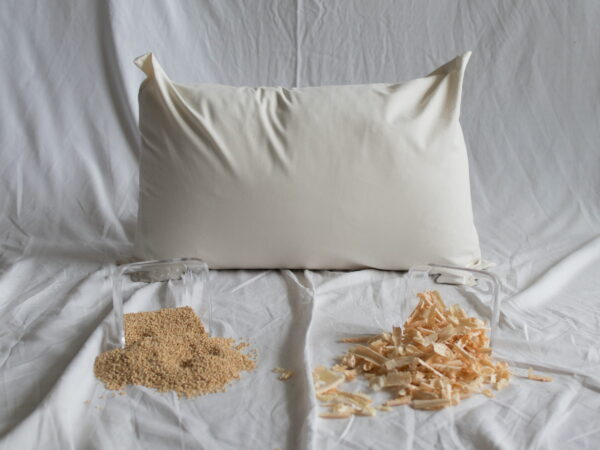 Hires-Zirbe-Kopfkissen von Kissenkorner auf weißem Bettlaken. Vor dem Kissen steht eine Schüssel mit Hirseschalen und Zirbenspänen.
