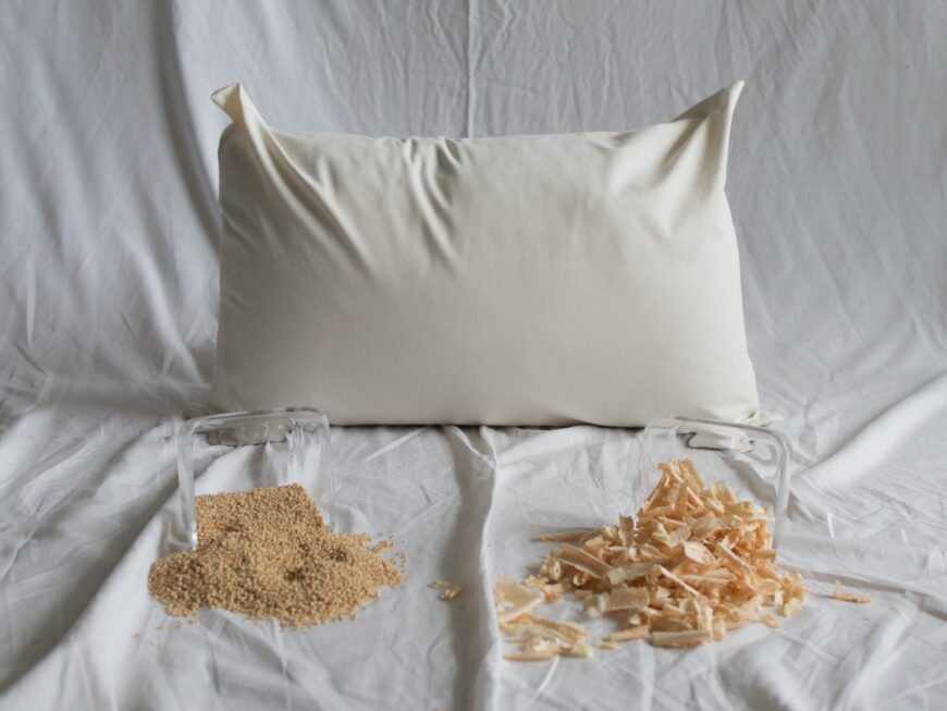 Hires-Zirbe-Kopfkissen von Kissenkorner auf weißem Bettlaken. Vor dem Kissen steht eine Schüssel mit Hirseschalen und Zirbenspänen.