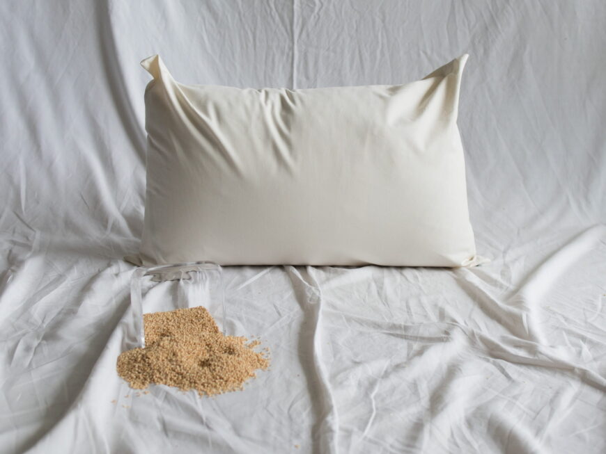 Hirsekopfkissen von Kissenkorner auf weißem Bettlaken. Vor dem Kissen steht eine Schüssel mit Hirseschalen.