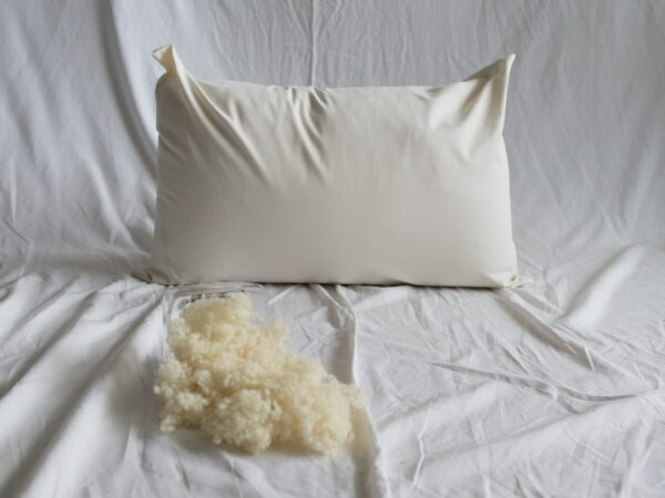 Schafwolle-Kopfkissen von Kissenkorner auf weißem Bettlaken. Vor dem Kissen steht eine Schüssel mit Schafwolle.