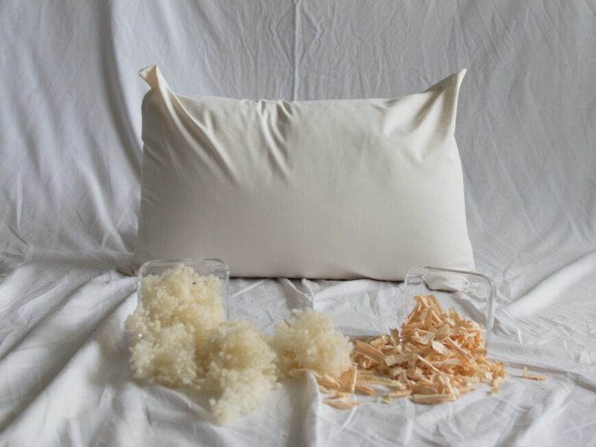 Schafwolle-Zirbe-Kopfkissen von Kissenkorner auf weißem Bettlaken. Vor dem Kissen steht eine Schüssel mit Schafwolle und Zirbenspänen.